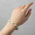 A close-up of a person's hand and wrist wearing a beaded bracelet made of alternating cream and mint green beads, set against a plain light gray background.