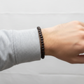 A beaded bracelet made of dark brown wooden beads, displayed on a light wood measuring block marked with '6in'.