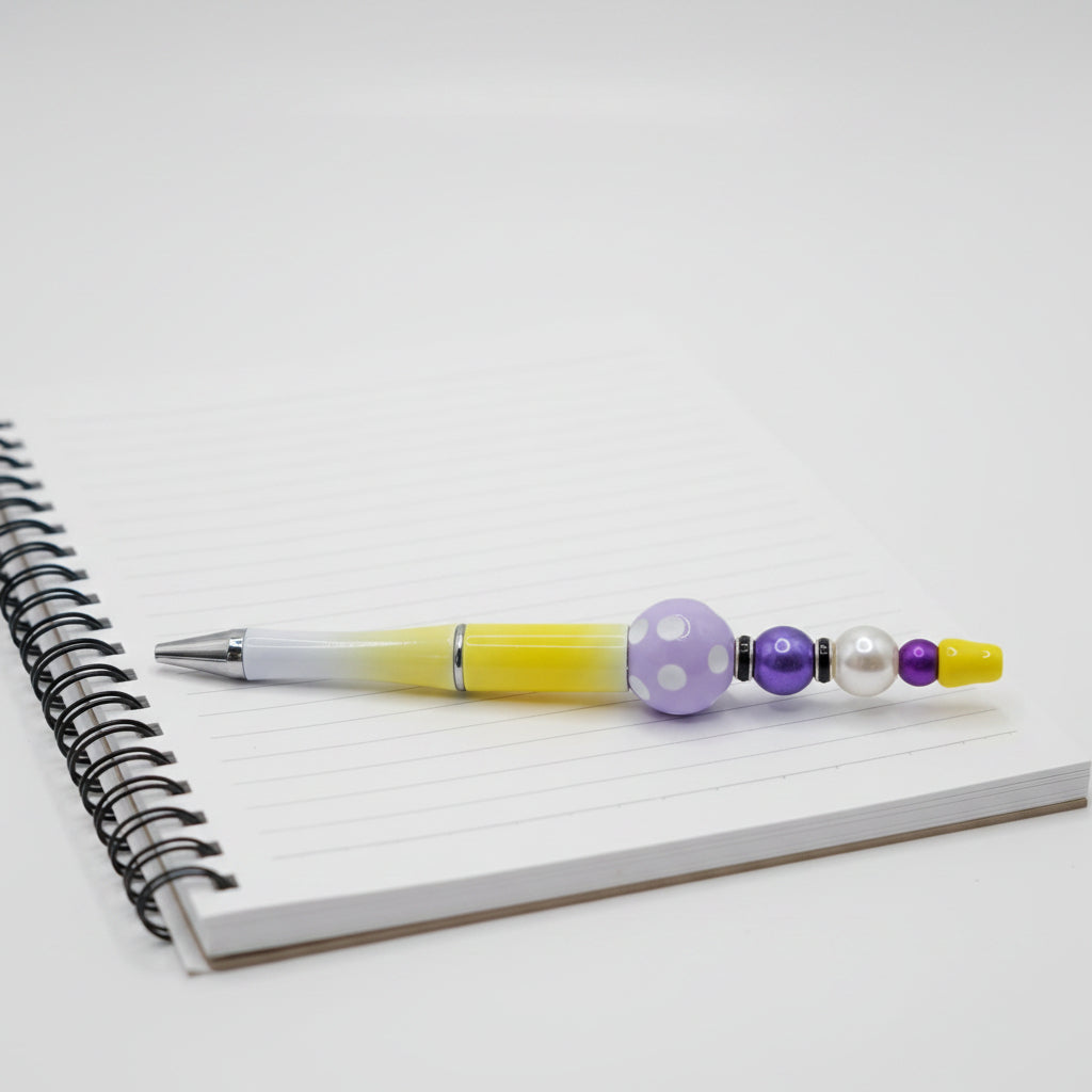 A decorative pen with a yellow-to-white gradient barrel, a purple beaded accent, and a silver tip, displayed on a wooden ruler with engraved size markings.