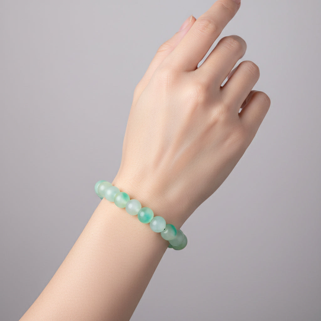 A beaded stretch bracelet made of round translucent jade-like beads in light green and aqua tones, displayed on a wooden measuring block marked 6in.