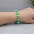 A beaded bracelet made of round turquoise-blue and yellow marbled beads, displayed on a wooden circular measuring stand marked 6 in.