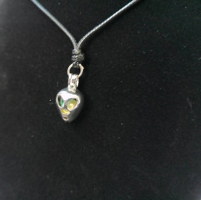 A silver pendant necklace featuring a stylized alien head charm with green and yellow eyes, suspended on a black cord against a black display bust.