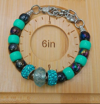 A handmade beaded bracelet with turquoise, dark purple, and black beads, featuring textured and glittery accents, displayed on a wooden measuring block marked 6 inches.