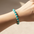 A beaded stretch bracelet with turquoise blue and white round beads worn on a person's wrist, shown in warm natural sunlight against a neutral beige background.