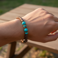 A beaded bracelet made of dark brown cylindrical beads with three turquoise blue round beads and small metallic spacer beads, displayed on a circular wooden measuring prop labeled 6in.