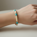 A close-up of a person's wrist wearing a beaded bracelet with turquoise blue beads, cream cylindrical beads, and small silver spacer beads.