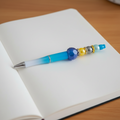 A decorative ballpoint pen with a blue and white gradient barrel, a faceted blue bead, yellow, gray, and light blue beads with gold spacer rings, placed on a wooden ruler over a light wood background.