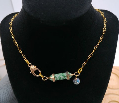 A gold chain necklace displayed on a black velvet jewelry bust. The necklace features a central cylindrical green stone bead with gold caps, a gold lobster clasp, and a small dangling blue crystal charm.