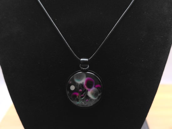 A black pendant necklace with a circular glass charm featuring purple, green, and white bubble-like patterns, displayed on a black velvet necklace bust.