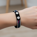 A black leather bracelet with silver-tone studs and a woven blue and red stripe detail, displayed on a light wood circular stand engraved with '6in'.