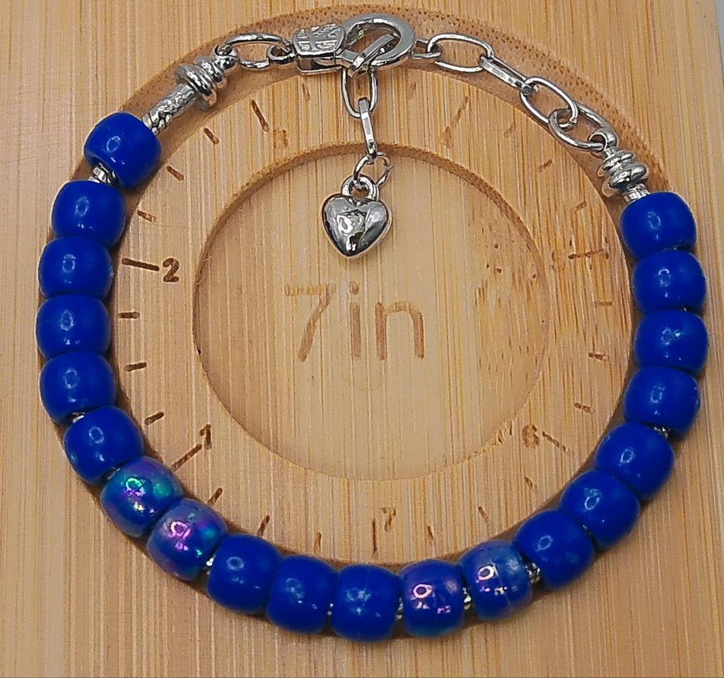 A beaded bracelet with glossy cobalt blue beads and silver-toned metal chain links, clasp, and heart charm, displayed on a light wood measuring block.