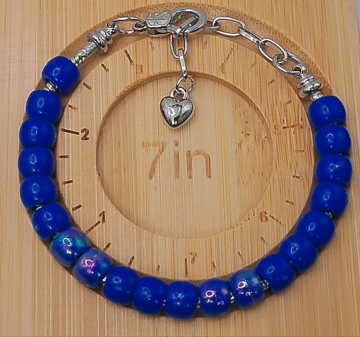 A beaded bracelet with glossy cobalt blue beads and silver-toned metal chain links, clasp, and heart charm, displayed on a light wood measuring block.