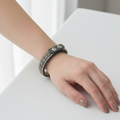 A dark gray leather cuff bracelet with a row of clear rhinestones on the front, displayed on a bamboo ring sizer stand marked 6in.