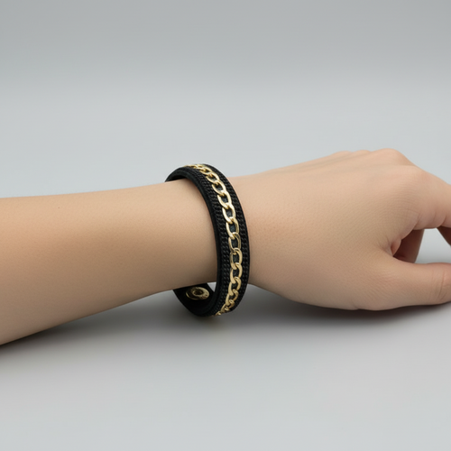 A black leather bracelet with a gold-tone chain link detail and gold snap buttons, displayed on a light wood grain surface.
