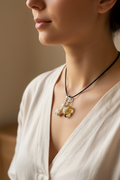 A close-up of a pendant necklace featuring a heart-shaped clear resin charm filled with yellow flower petals and a small tree, alongside four white shell beads on a black cord, set against a black background.