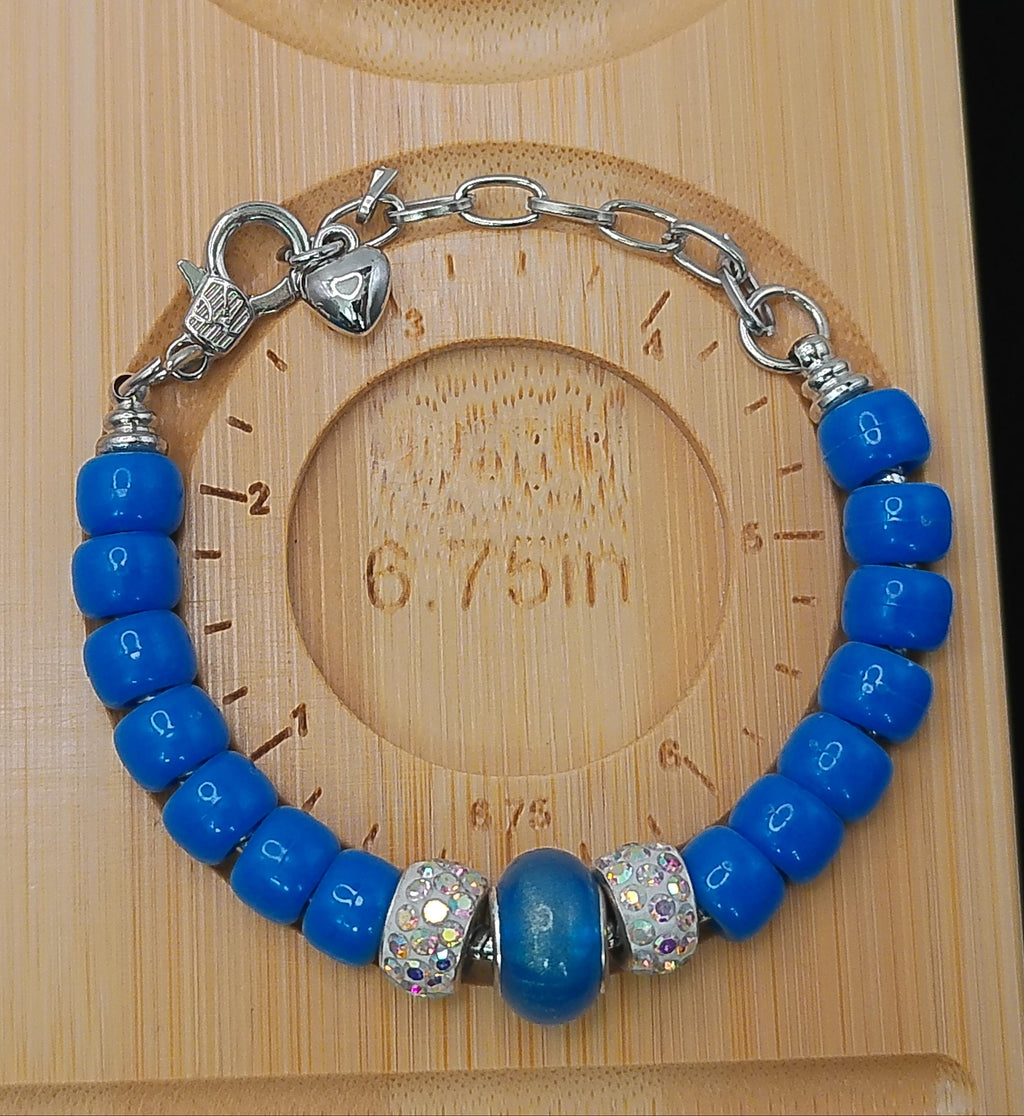 A blue beaded bracelet with silver-toned chain links, a heart-shaped clasp, and two decorative silver spacer beads, displayed on a wooden measuring board.