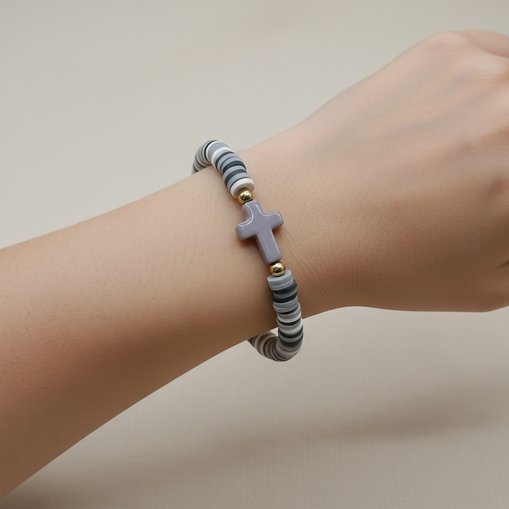 A beaded bracelet with alternating black, gray, and white stripes, featuring a central lavender-purple cross charm and small gold spacer beads, displayed on a wooden stand.