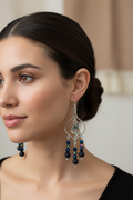 A pair of silver-toned dangle earrings with rose charms and dark blue and black beads, displayed on a tan card over a light wood background.