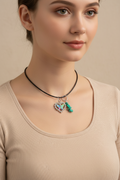 A close-up of a necklace with a heart-shaped butterfly charm outlined in clear rhinestones and a separate green stone charm, both hanging from silver rings on a dark cord against a black background.