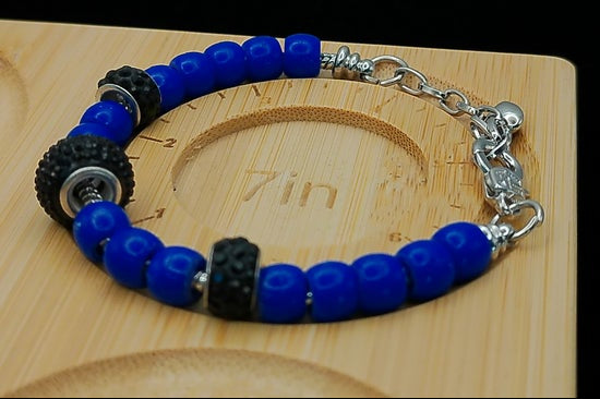 A beaded bracelet with bright blue round beads, two black textured spacer beads, and silver-toned chain links and clasp, displayed on a light wood block.