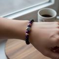 A beaded stretch bracelet displayed on a wooden measuring block. The bracelet features matte black round beads, three glossy purple beads, two light beige beads, and a central silver lion head charm.
