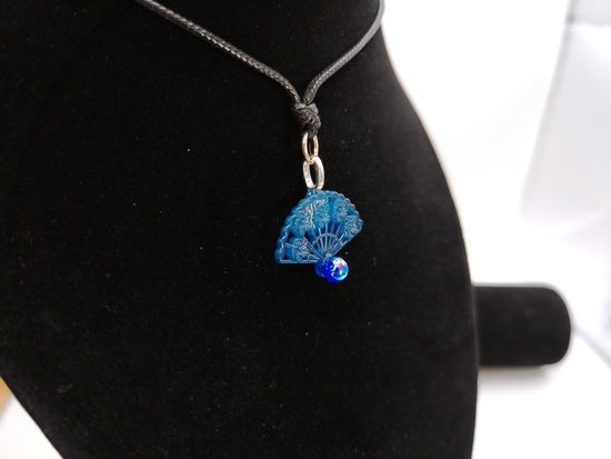 A blue fan-shaped pendant necklace with a small blue bead charm and silver-toned metal, displayed on a black velvet jewelry bust.