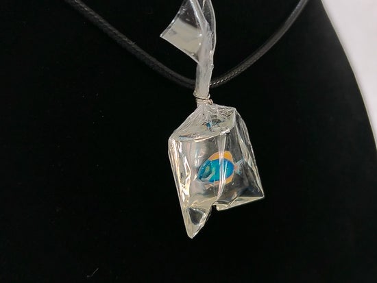 A clear faceted glass or resin pendant shaped like a small bag, containing a small blue and orange fish, hanging on a black cord against a black display background.
