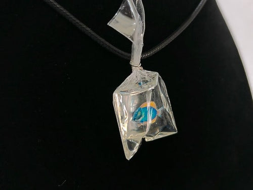 A clear faceted glass or resin pendant shaped like a small bag, containing a small blue and orange fish, hanging on a black cord against a black display background.